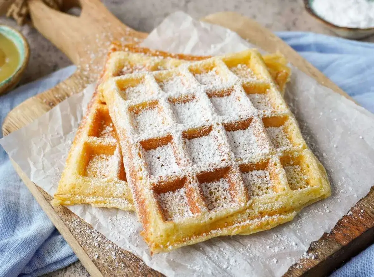Zwei fluffige Waffeln, best&auml;ubt mit Puderzucker, auf einem Holzbrett. Ein perfektes Rezept f&uuml;r fluffige Waffeln!