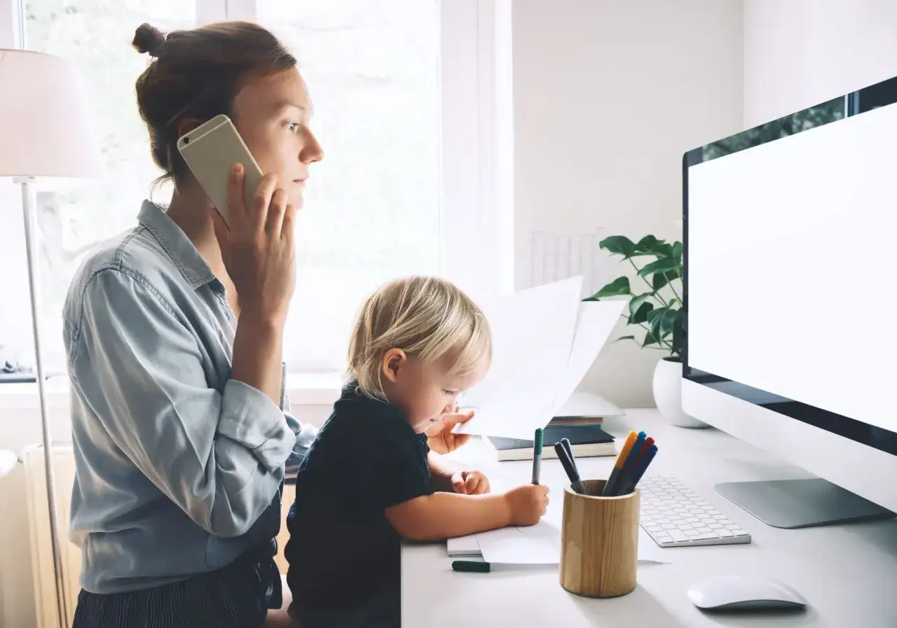 Kobieta na urlopie opiekuńczym rozmawia przez telefon, podczas gdy jej dziecko rysuje przy biurku.