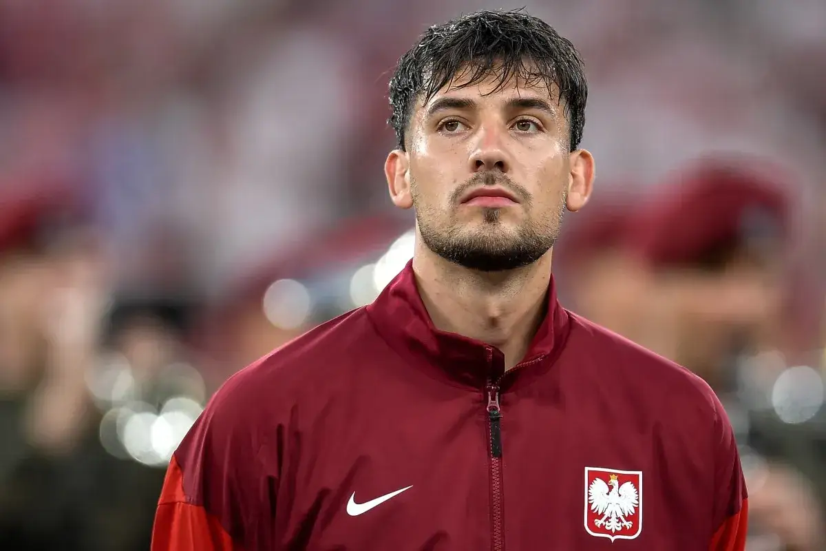 Piłkarz w bordowej kurtce z orłem, zamyślony, być może po trudnym meczu lub zmagający się z **moder kontuzja**.