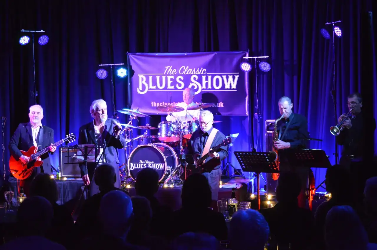 Zesp&oacute;ł gra blues na żywo. Na scenie gitarzysta, saksofonista, trębacz i perkusista. Baner głosi 