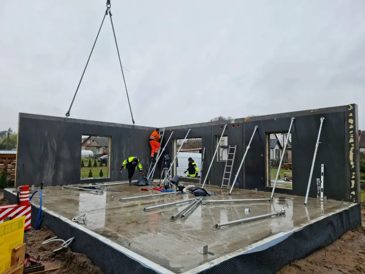 Budowa domu z gotowych elementów betonowych. Dźwig podnosi ścianę, pracownicy na drabinach mocują ją do fundamentów.