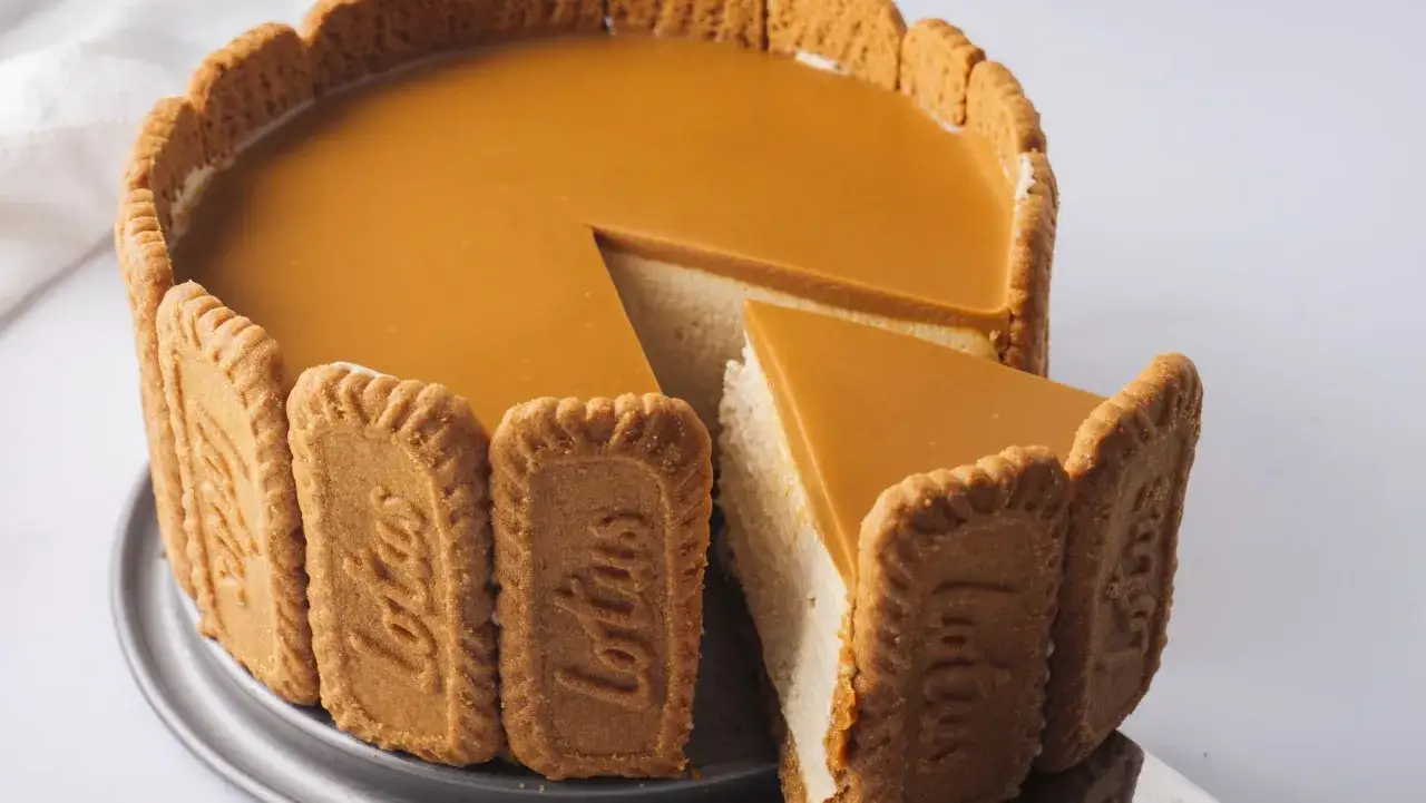 Ein köstlicher Lotus Cheesecake, umhüllt von knusprigen Lotus Keksen, mit einer glatten Karamellglasur. Ein Stück ist bereits angeschnitten.