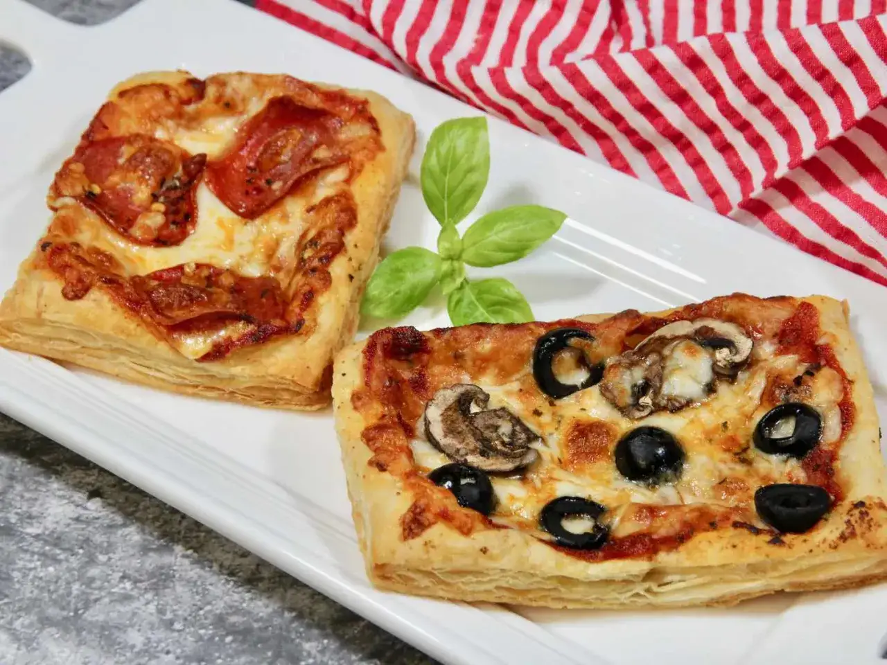 Dwie pyszne pizze z ciasta francuskiego: jedna z salami, druga z oliwkami i pieczarkami. Idealne na szybką przekąskę.