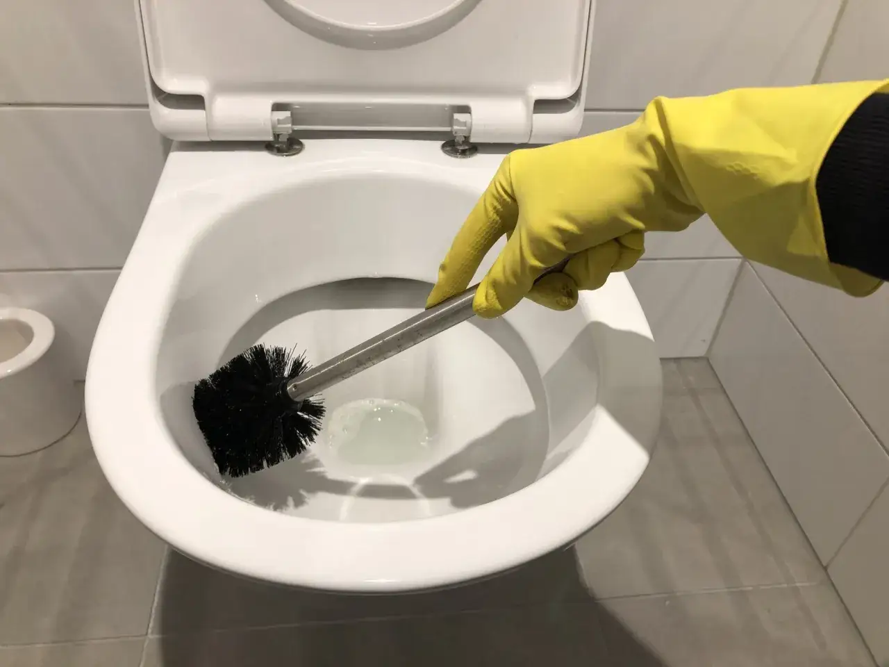 Hand in gele handschoen maakt de wc vloer schoon met een borstel.