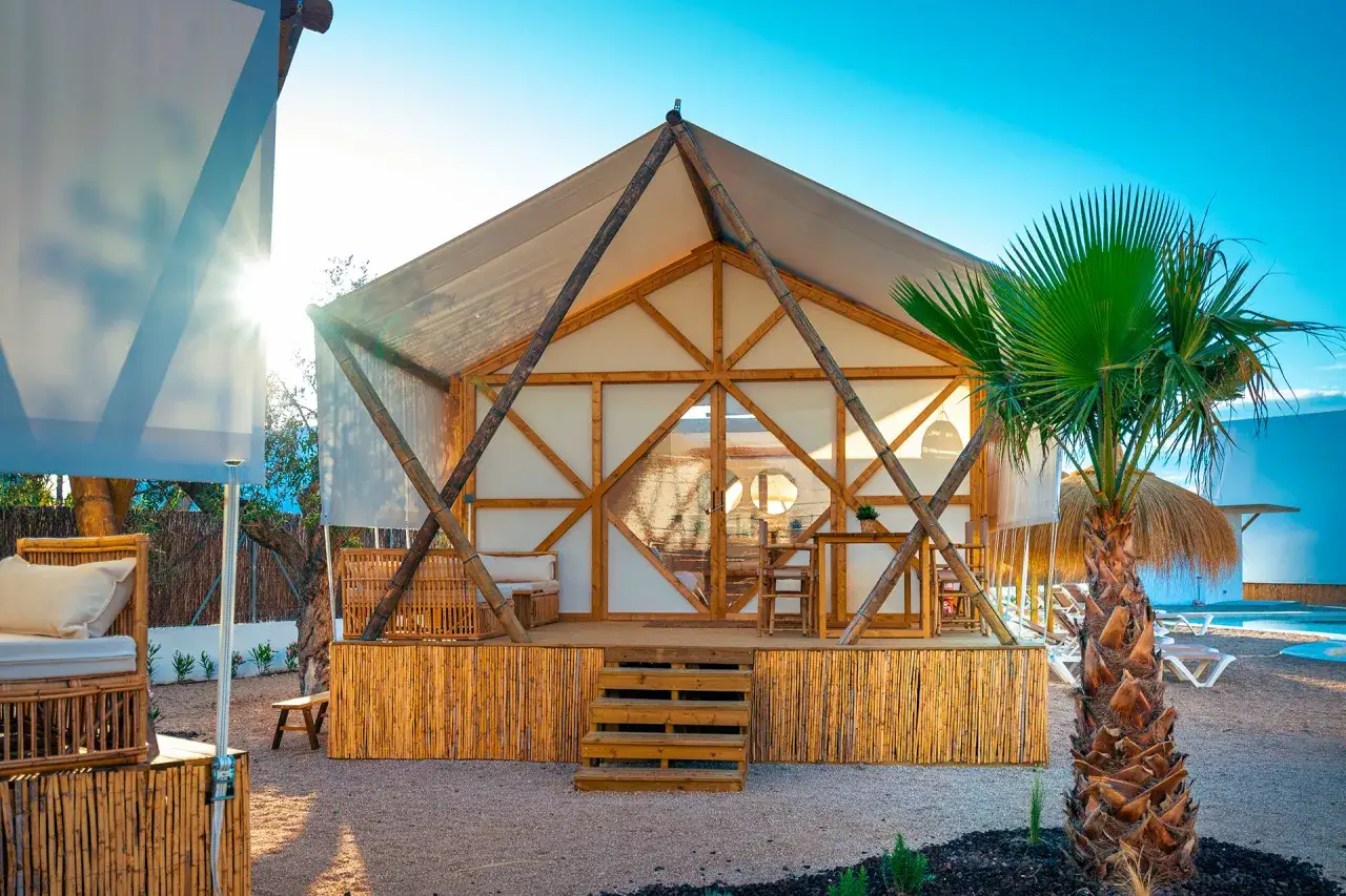 Glamping para niños Comunidad Valenciana: tienda de bambú con muebles de exterior y palmera bajo el sol.