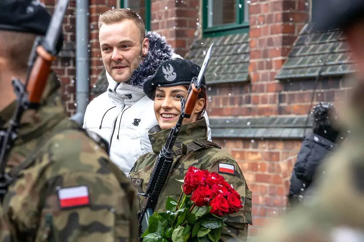 Uśmiechnięta żołnierka z bukietem czerwonych róż, gotowa do złożenia przysięgi wojskowej. Obok niej stoi mężczyzna w białej kurtce.