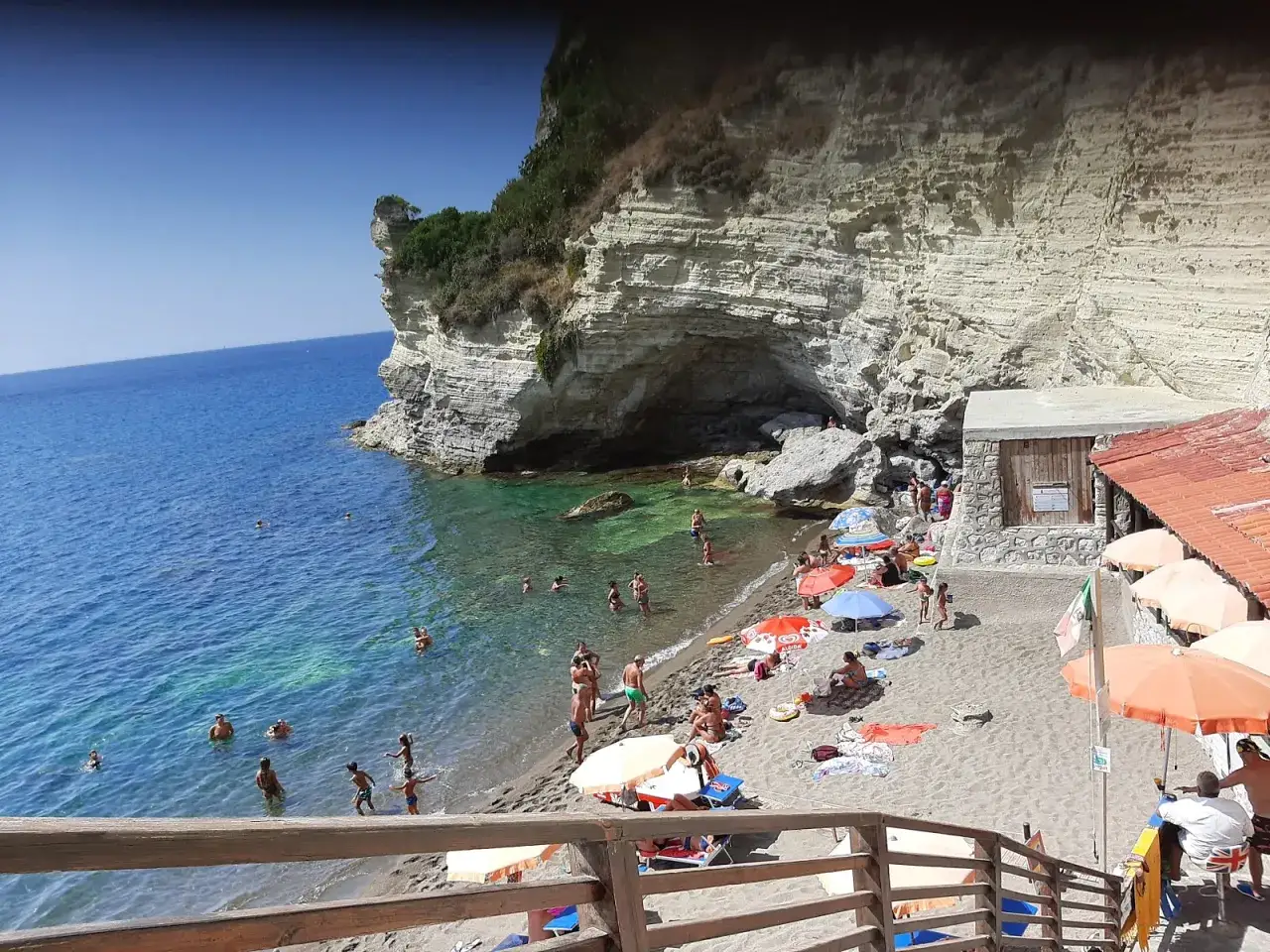 Spiaggia affollata con scogliera imponente, bagnanti nell'acqua cristallina e ombrelloni colorati. La **cava grado Ischia** offre un rifugio naturale.