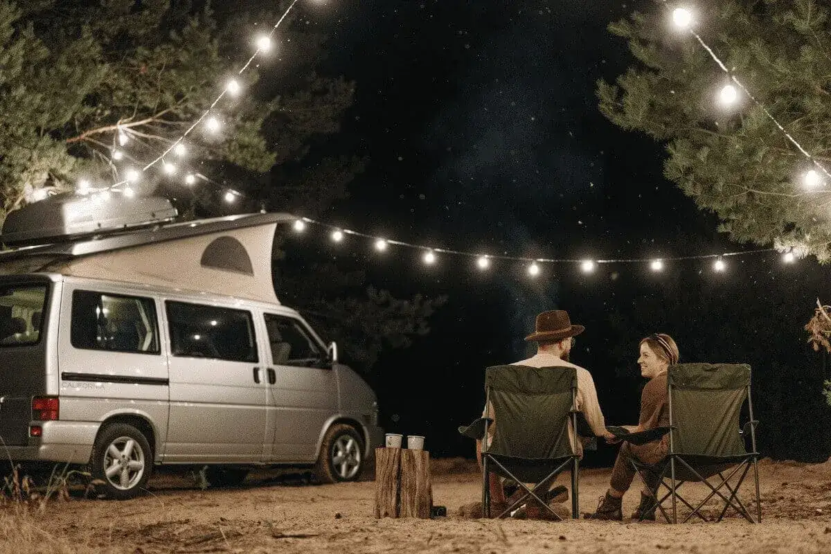 Pareja acampando bajo luces de feria en la costa atlántica francesa. Una furgoneta camper y sillas plegables crean un ambiente acogedor.