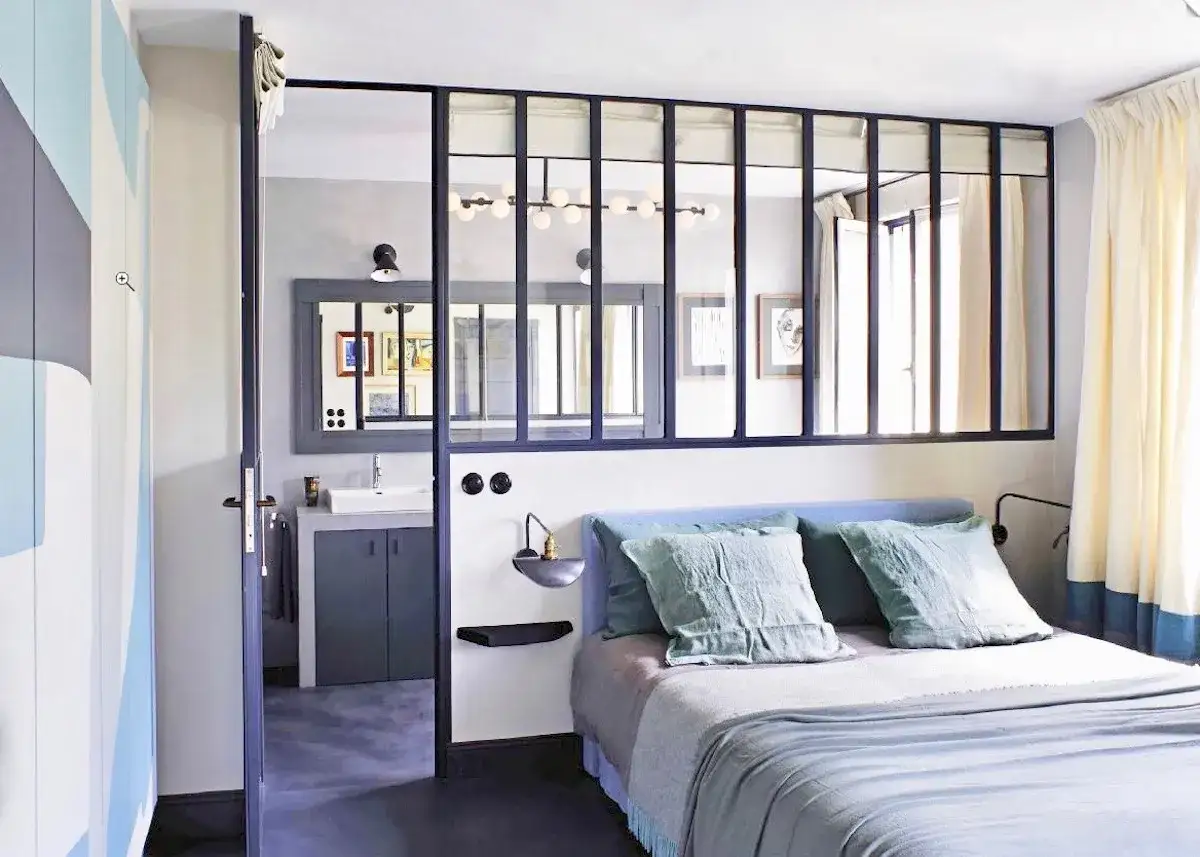 Chambre parentale avec salle de bain ouverte, s&eacute;par&eacute;e par une verri&egrave;re. Lit bleu, coussins verts, rideaux clairs.