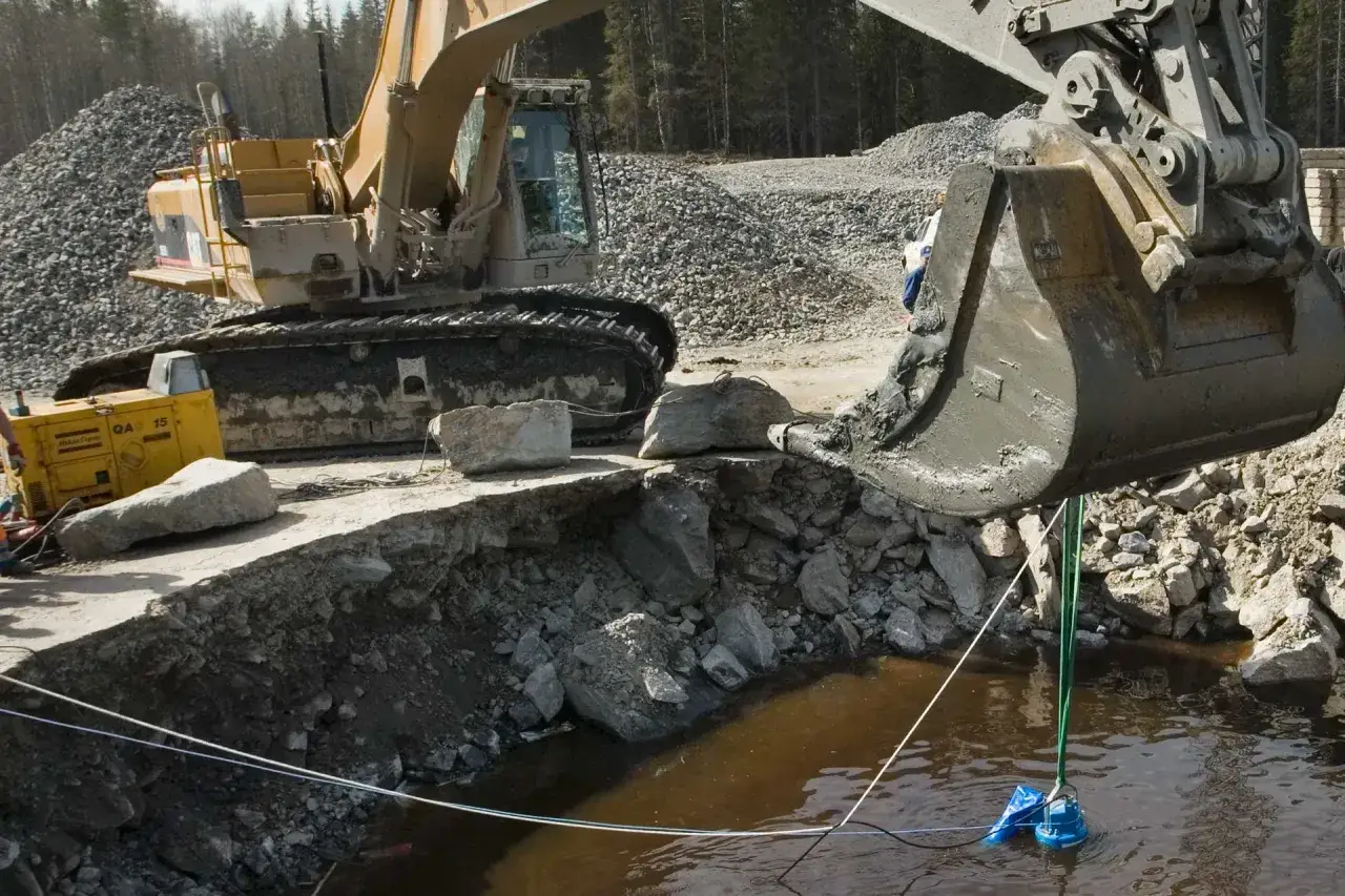 Excavadora trabajando cerca de una motobomba sumergida en agua turbia.