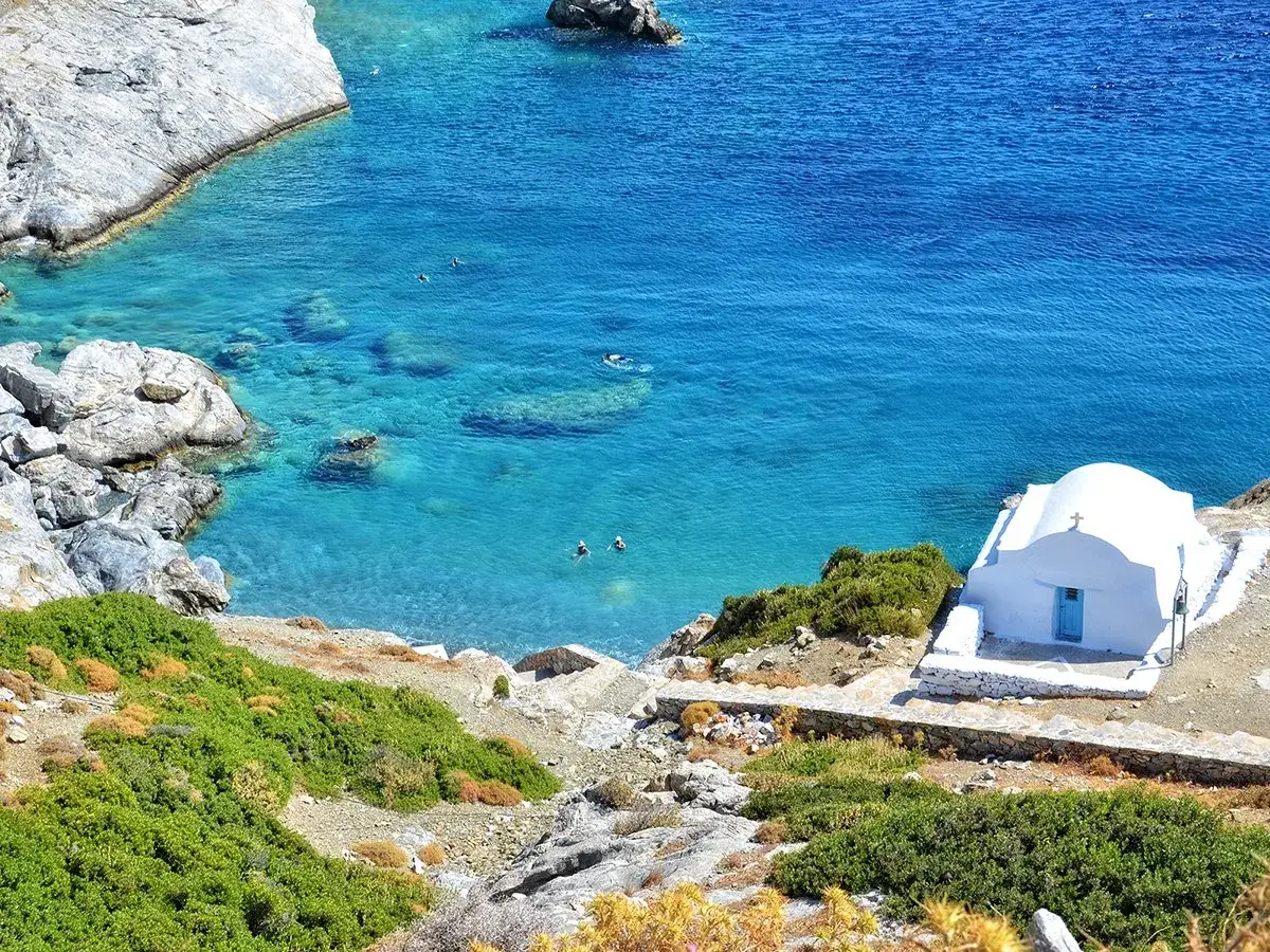 En vit kyrka med bl&aring; d&ouml;rr vid en turkos vik p&aring; Amorgos, Grekland. N&aring;gra badar i det klara vattnet.