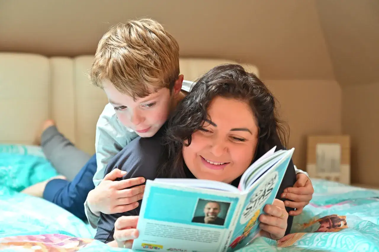 Mama i syn leżą na łóżku, czytając książki dla par. Chłopiec przytula się do mamy, oboje z uśmiechem na twarzy.