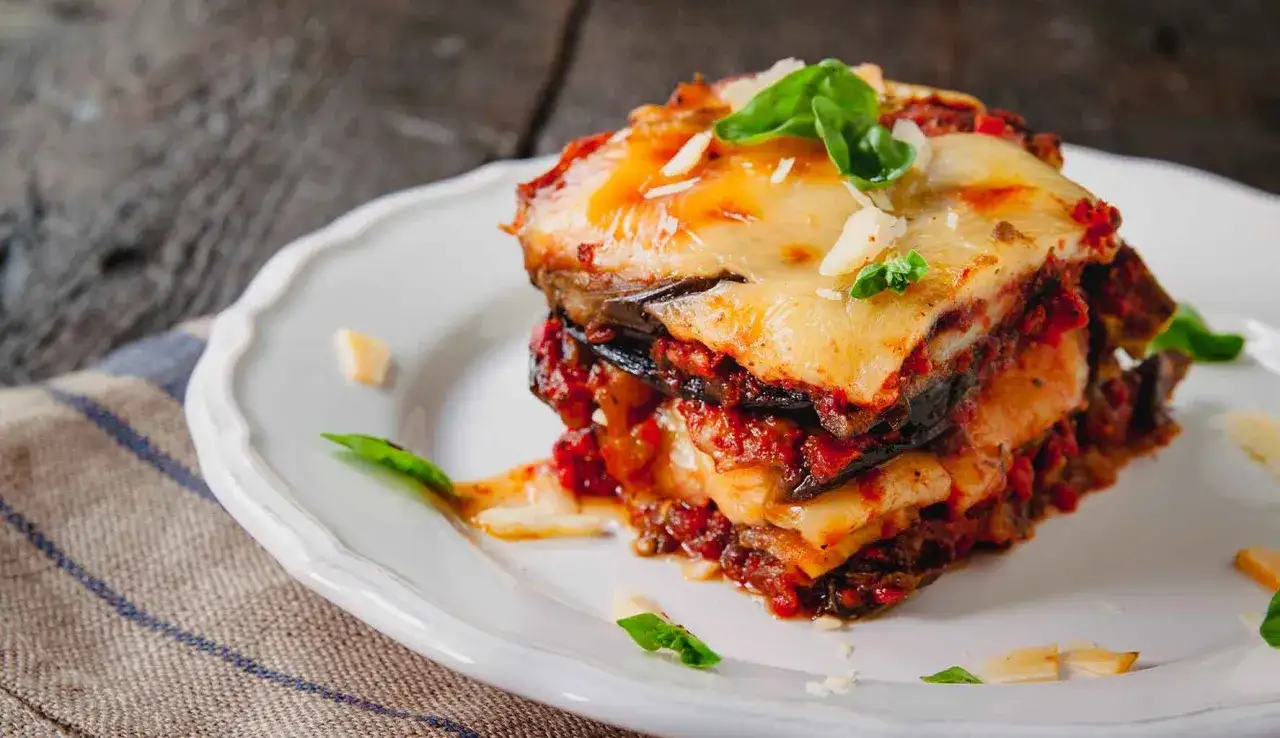 Una fetta di parmigiana di melanzane non fritte, con strati di melanzane, sugo e formaggio filante, guarnita con basilico fresco.