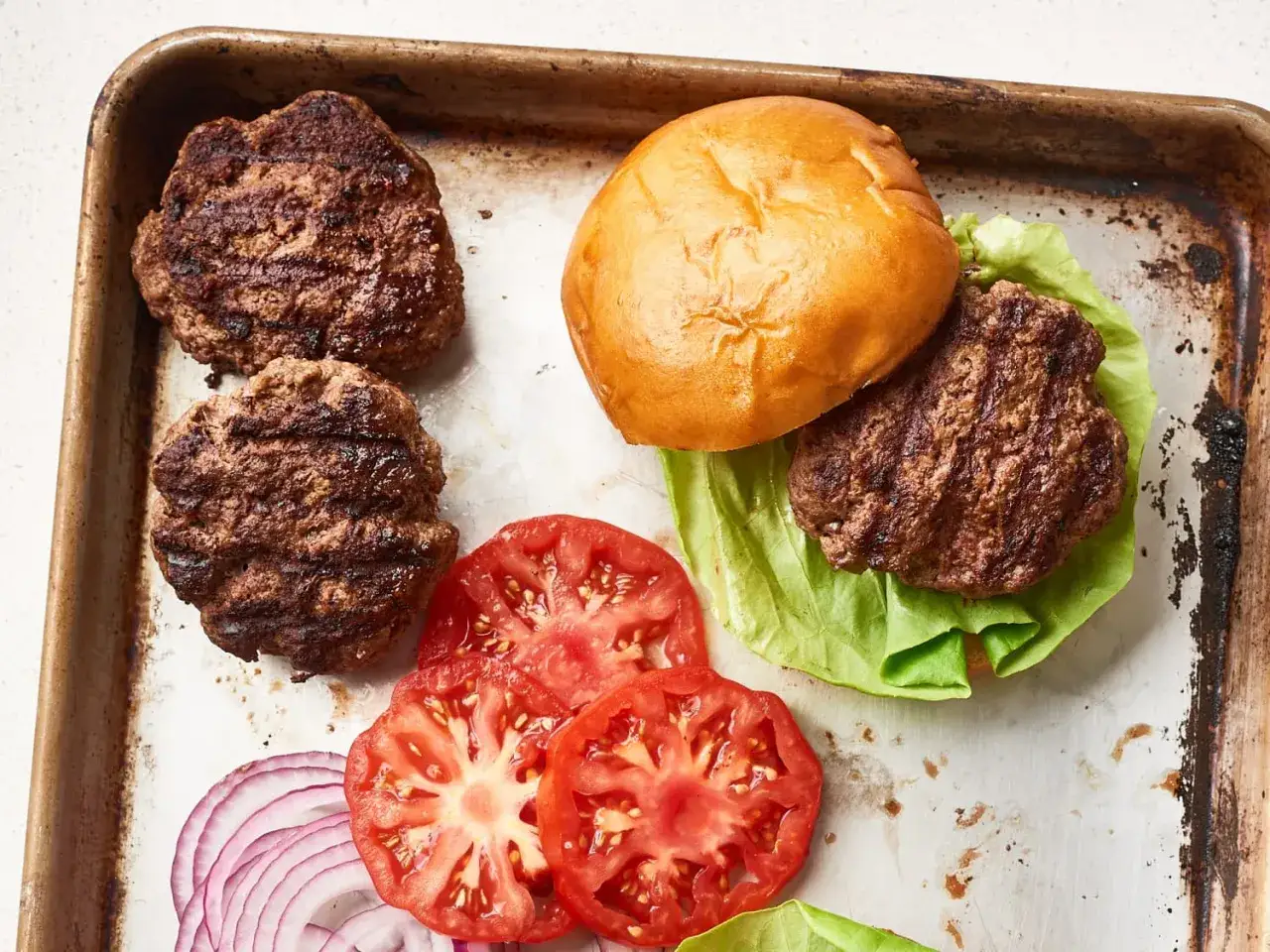 Grillowane mięso na burgery, bułka, sałata, pomidor i cebula na blasze. Idealne składniki na pysznego burgera.