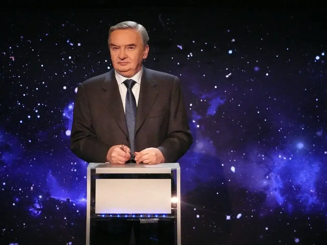 Mężczyzna w garniturze stoi przy mównicy na tle kosmicznego nieba. Jeden z dziesięciu od kiedy zaczął karierę.