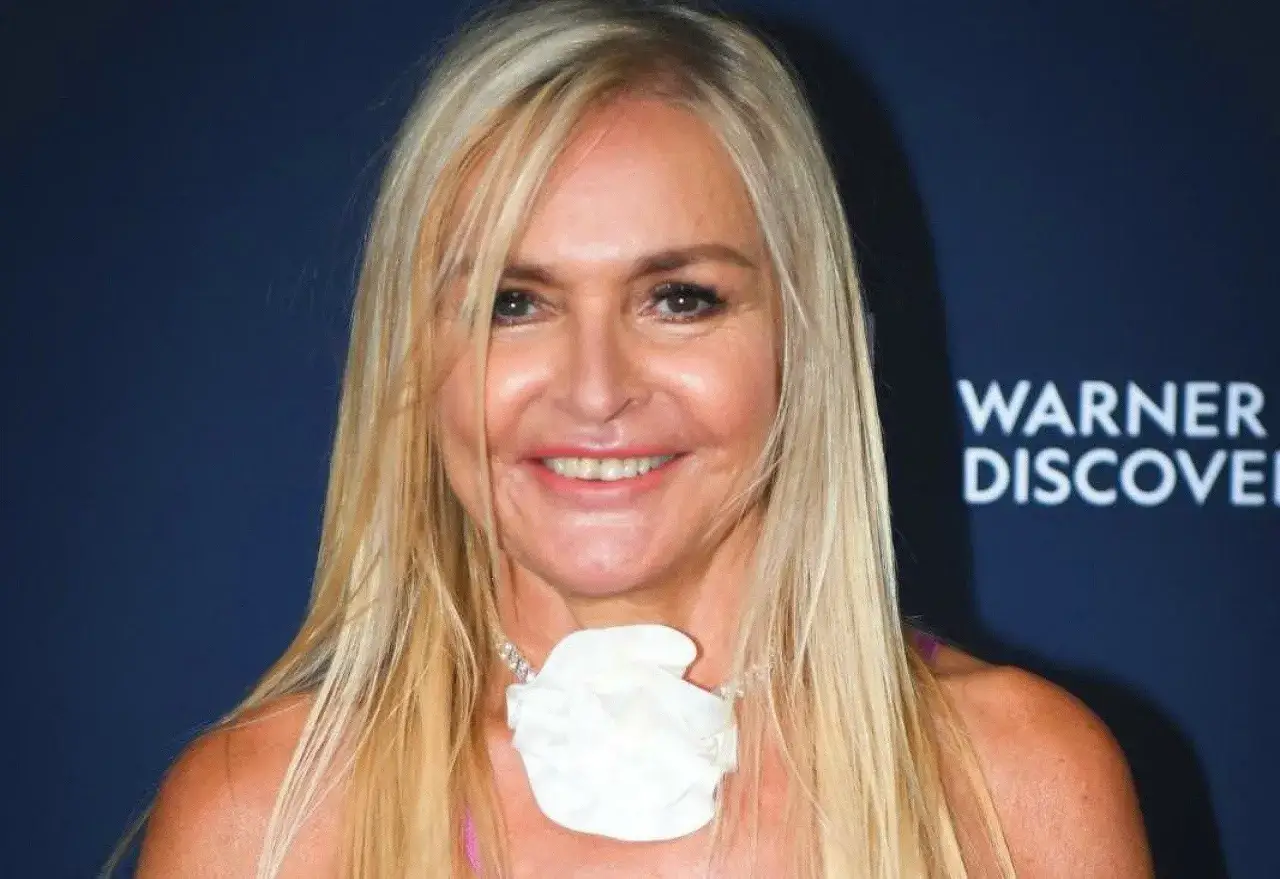 Uśmiechnięta blondynka z białym kwiatem na szyi. W tle logo Warner Discovery. Wiele osób zastanawia się, ile lat ma Monika Olejnik.