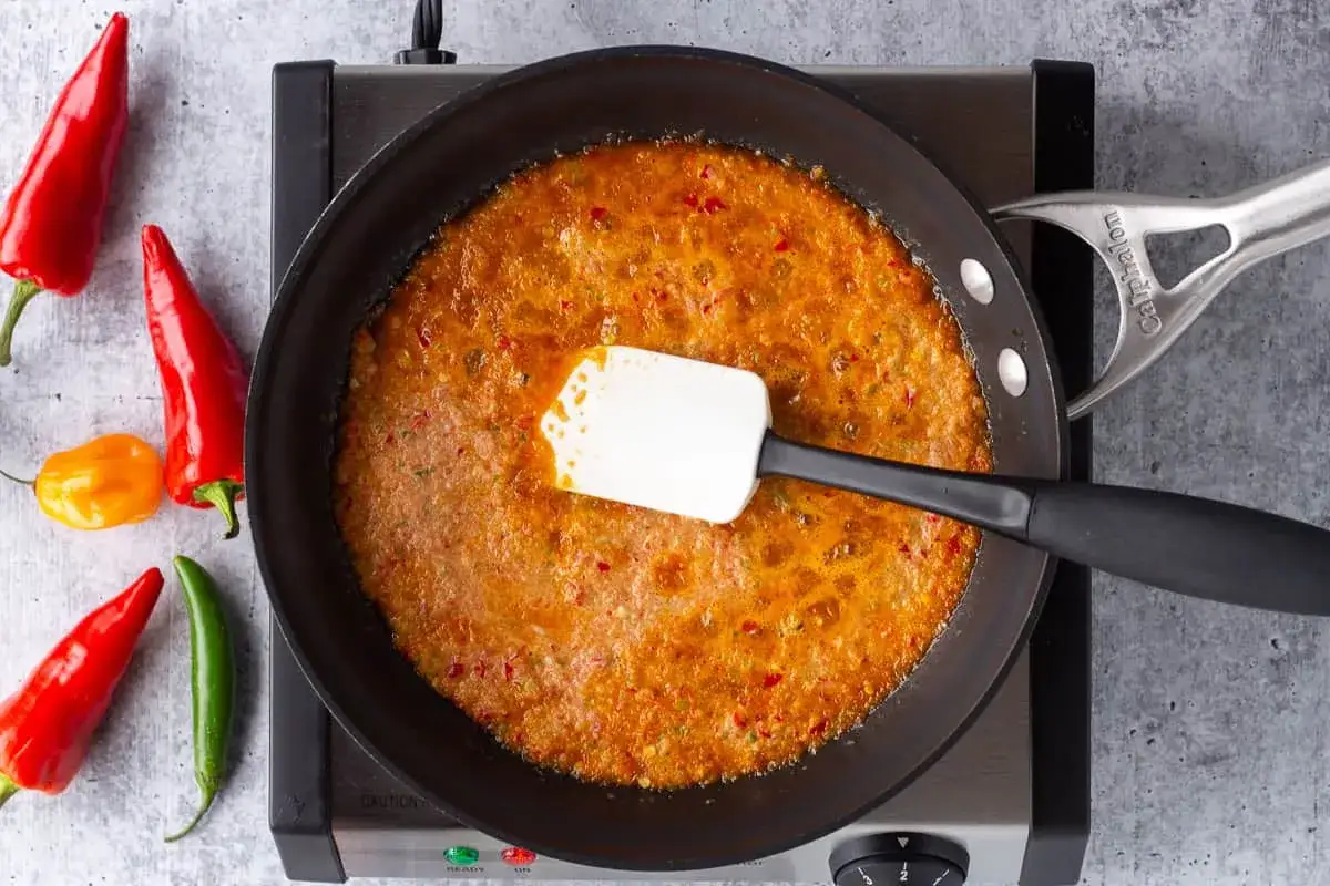 En panna med bubblande sambal oelek sås, omgiven av färska röda och gröna chilifrukter. En spatel rör om i den kryddiga såsen.