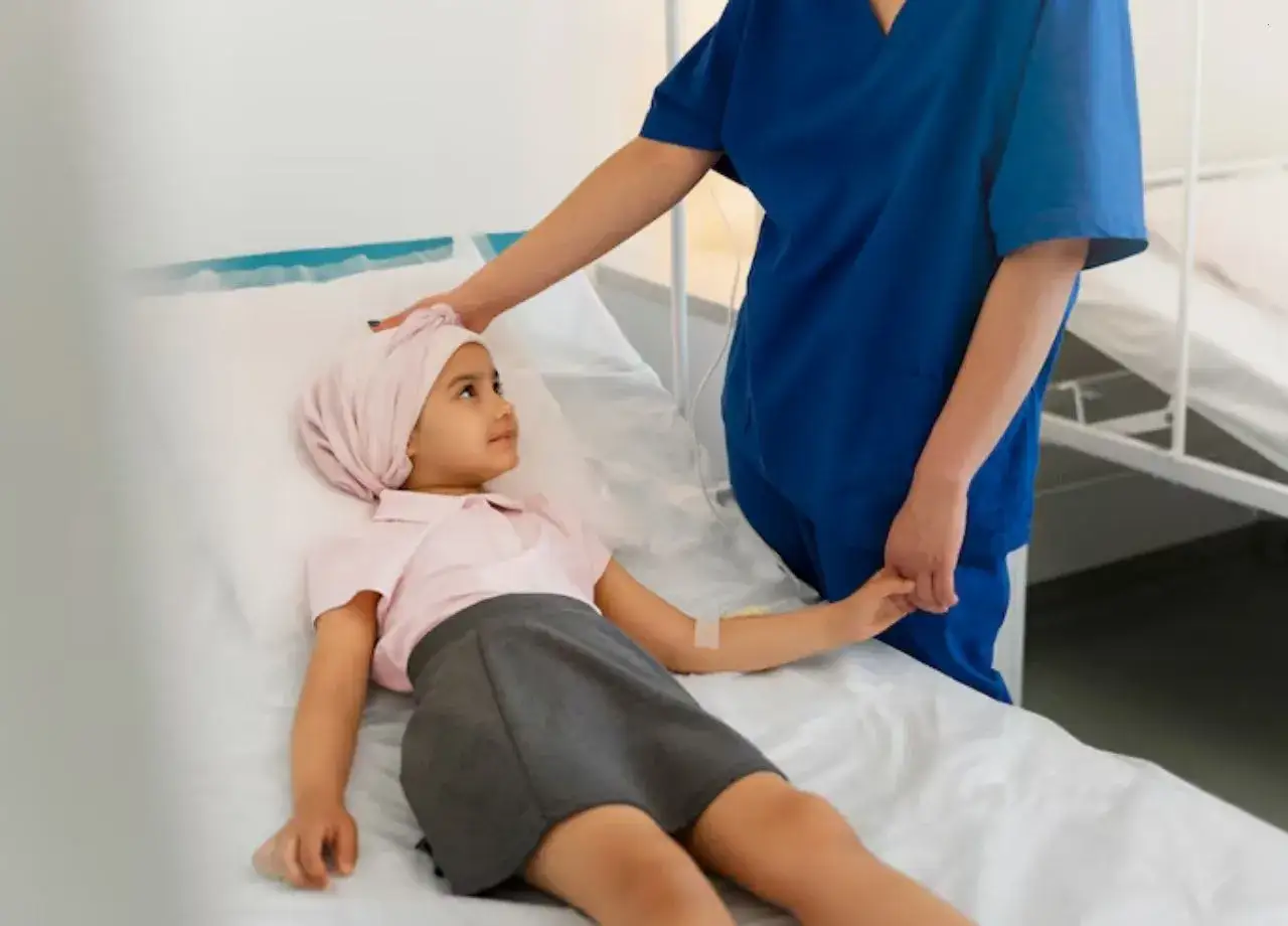 Mała pacjentka w szpitalnym łóżku, pielęgniarka kładzie jej rękę na głowie. Dziecko ma założony plaster z wenflonem, co sugeruje podawanie leków, być może znieczulenie miejscowe.
