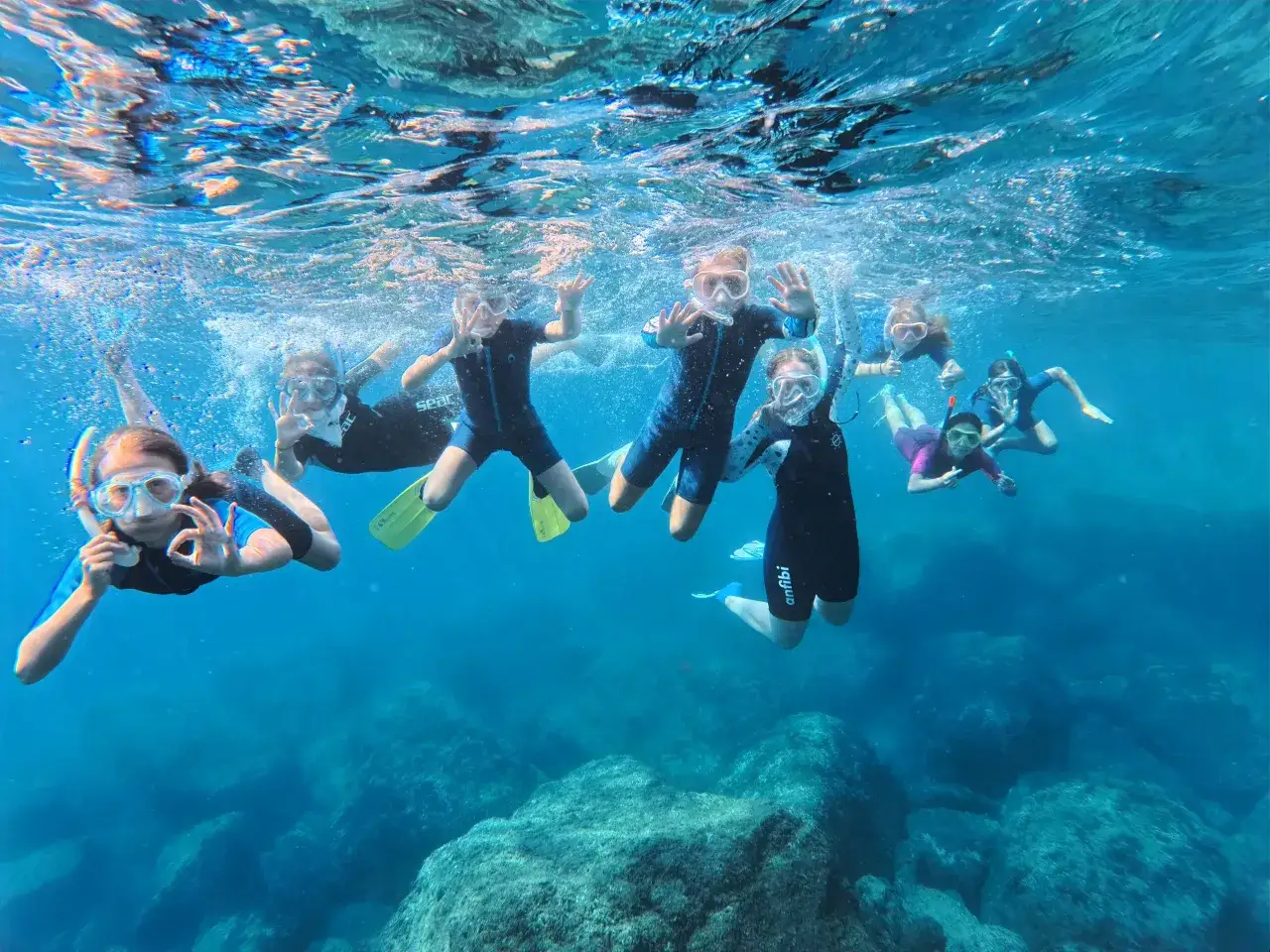 Bambini felici fanno snorkeling in un'area marina protetta, salutando con le mani tra rocce e alghe.