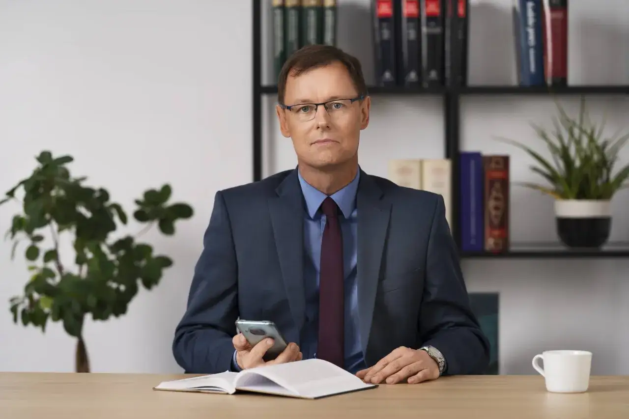 Mężczyzna w garniturze siedzi przy biurku z telefonem i otwartą książką.