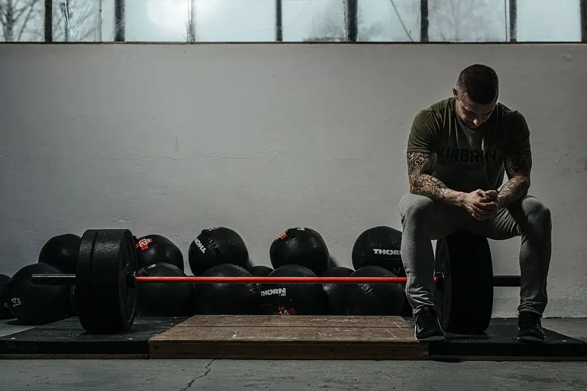 Mężczyzna z tatuażami siedzi na talerzu od sztangi, otoczony piłkami lekarskimi i gryfem. Inspiracja, jak ćwiczyć crossfit w domu.