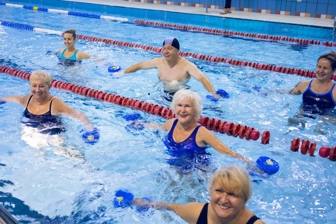Grupa osób w różnym wieku cieszy się ćwiczeniami w wodzie, używając niebieskich przyrządów do aqua aerobiku.