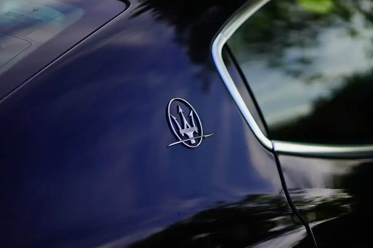 Błyszczący, chromowany znaczek Maserati z tr&oacute;jzębem Neptuna na ciemnoniebieskim lakierze samochodu.