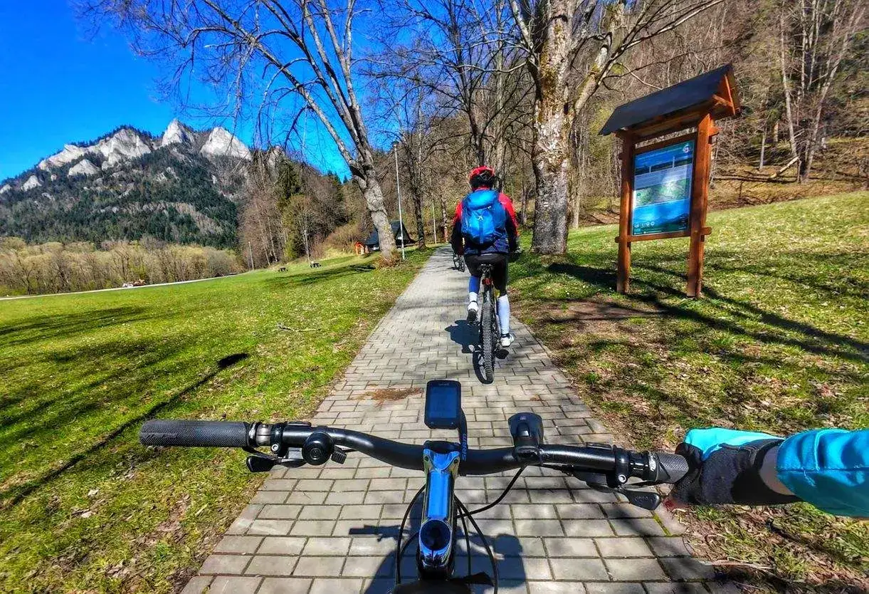 Wyprawa rowerem w góry. Dwie osoby na rowerach jadą ścieżką pośród drzew, z widokiem na skaliste szczyty.