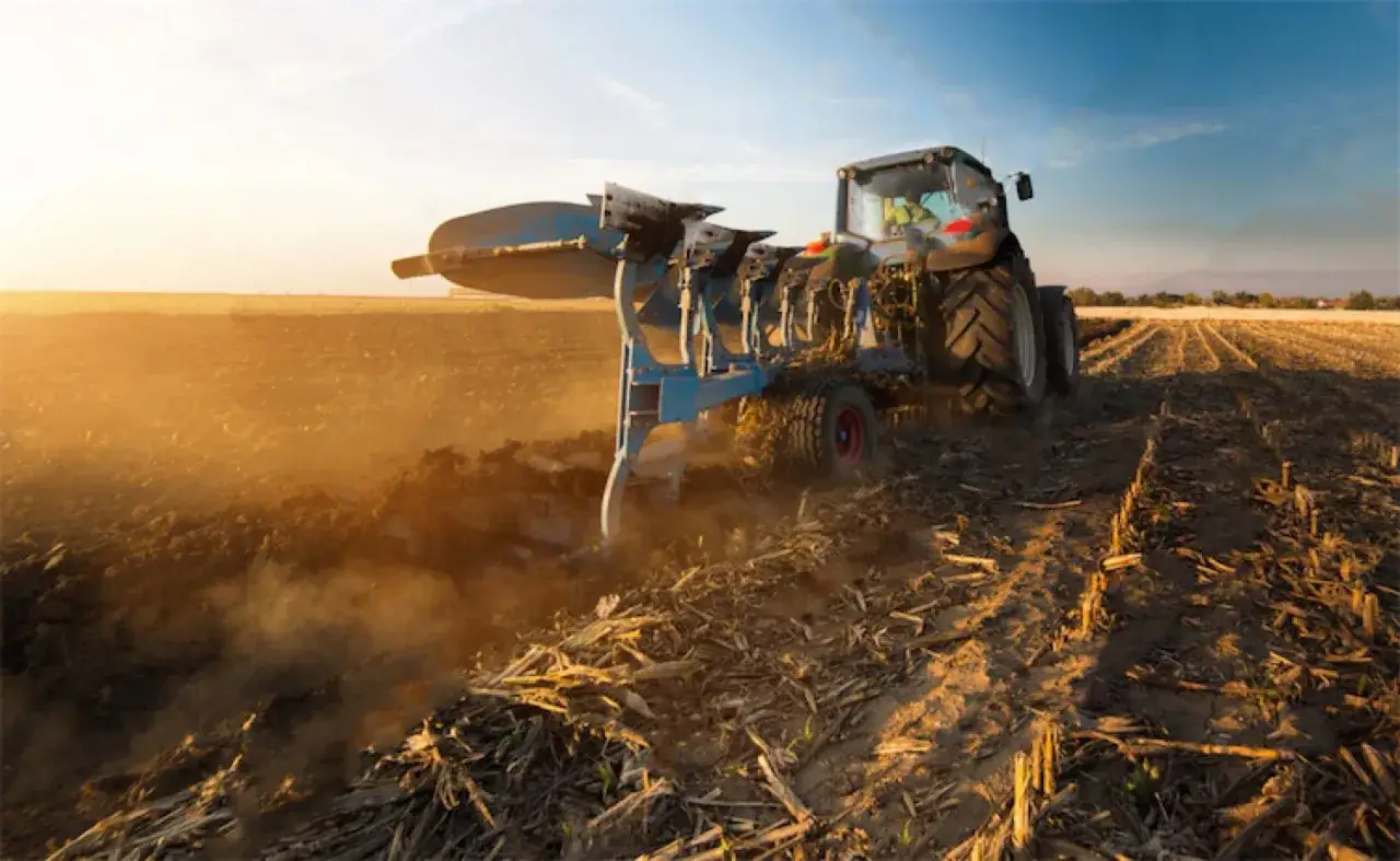 Traktor z pługiem wykonuje podorywkę na polu, wzbijając tumany kurzu w złotym świetle zachodzącego słońca.