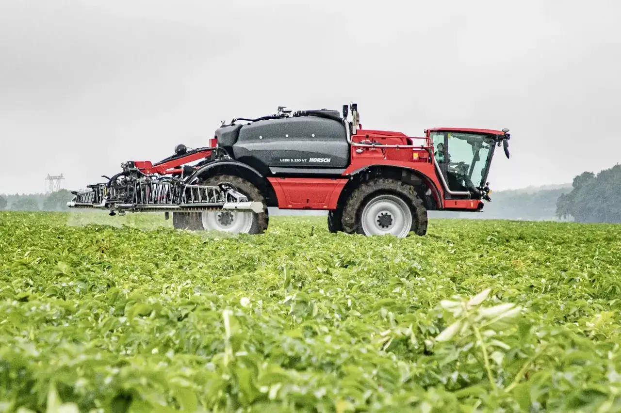 Czerwony opryskiwacz polowy HORSCH Leer S.230 VT w akcji, rozprowadzający środek ochrony roślin, np. metrybuzynę, na zielonym polu.