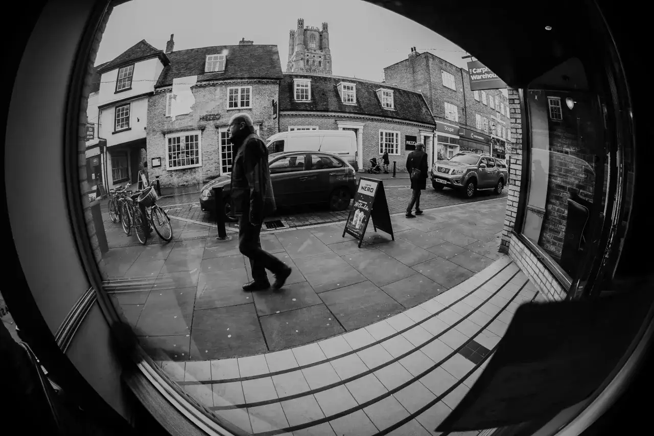 Calle con edificios, coches y gente. Un hombre camina, visto con ojo de pez.