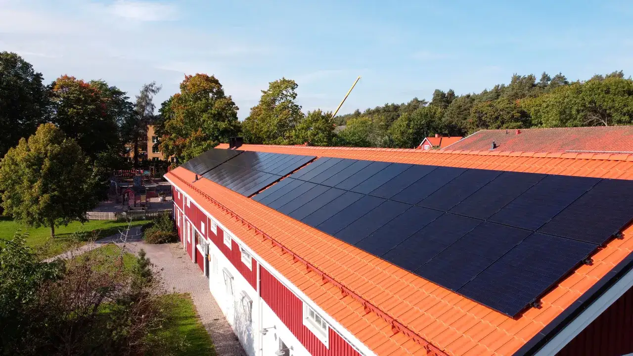 Solceller p&aring; taket till en r&ouml;d byggnad, troligen en del av ett solceller brf-projekt. Gr&ouml;nskande tr&auml;d och en lekplats syns i bakgrunden.