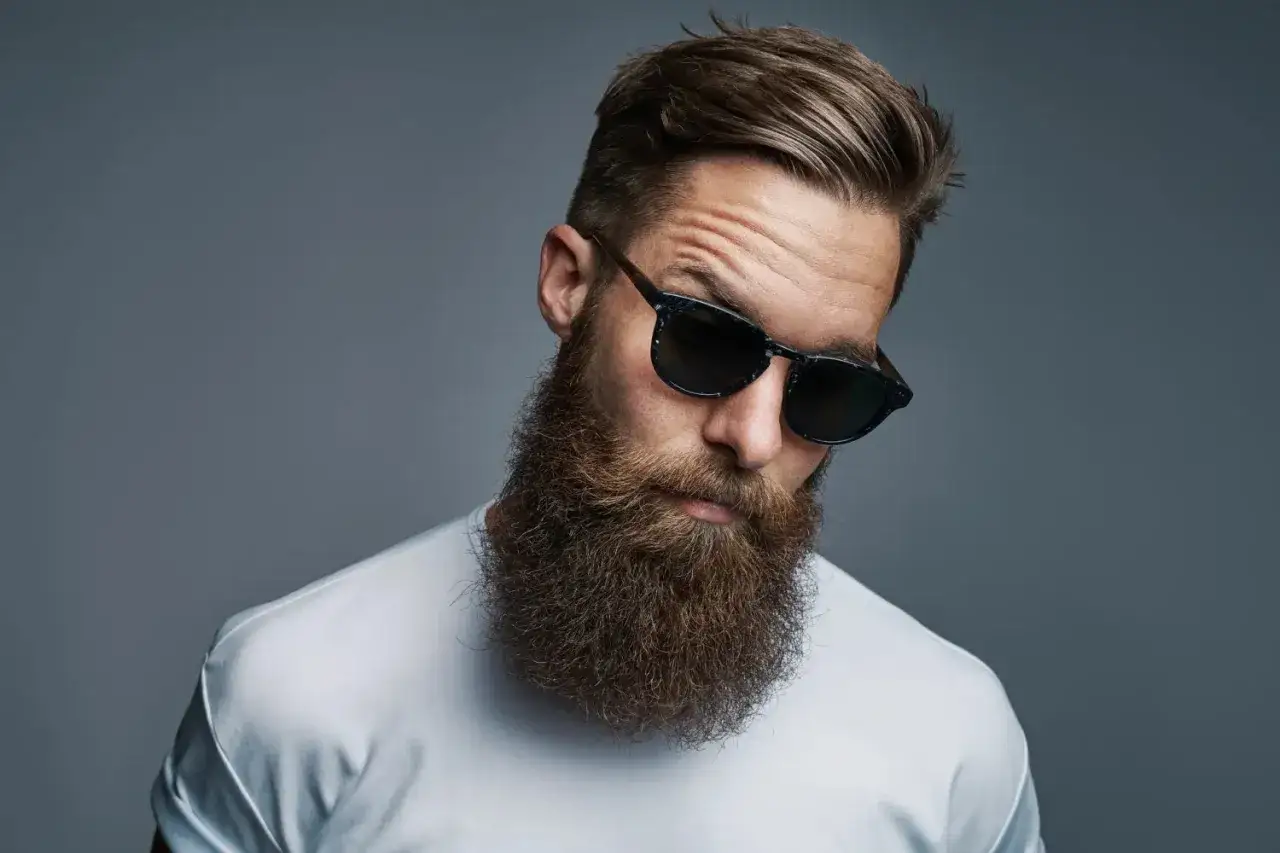 Hombre con barba hipster corta, gafas de sol oscuras y camiseta blanca, mirando hacia abajo con una expresi&oacute;n seria.