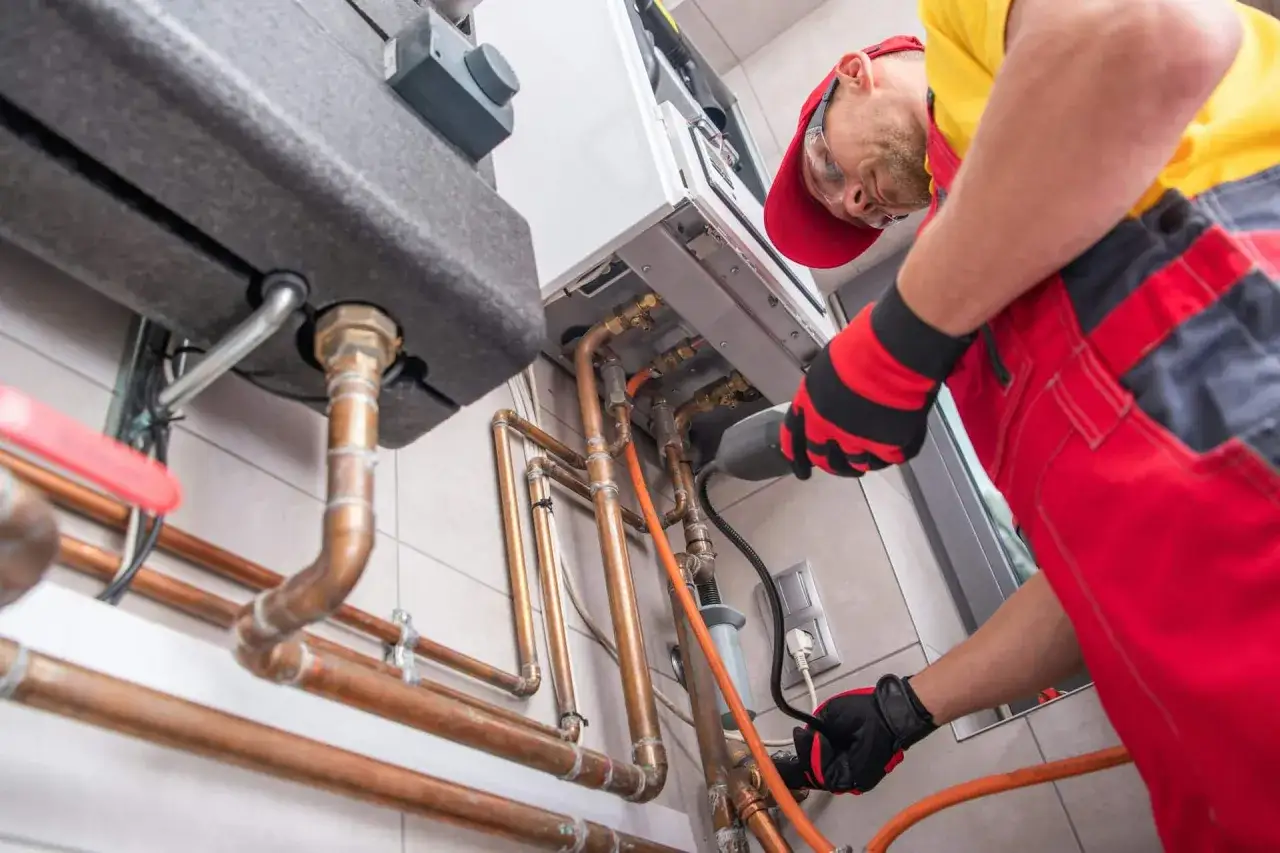 Un technicien en combinaison rouge et jaune travaille sur une chaudière à gaz. Il est en train de réaliser une intervention d'aide remplacement chaudière gaz.