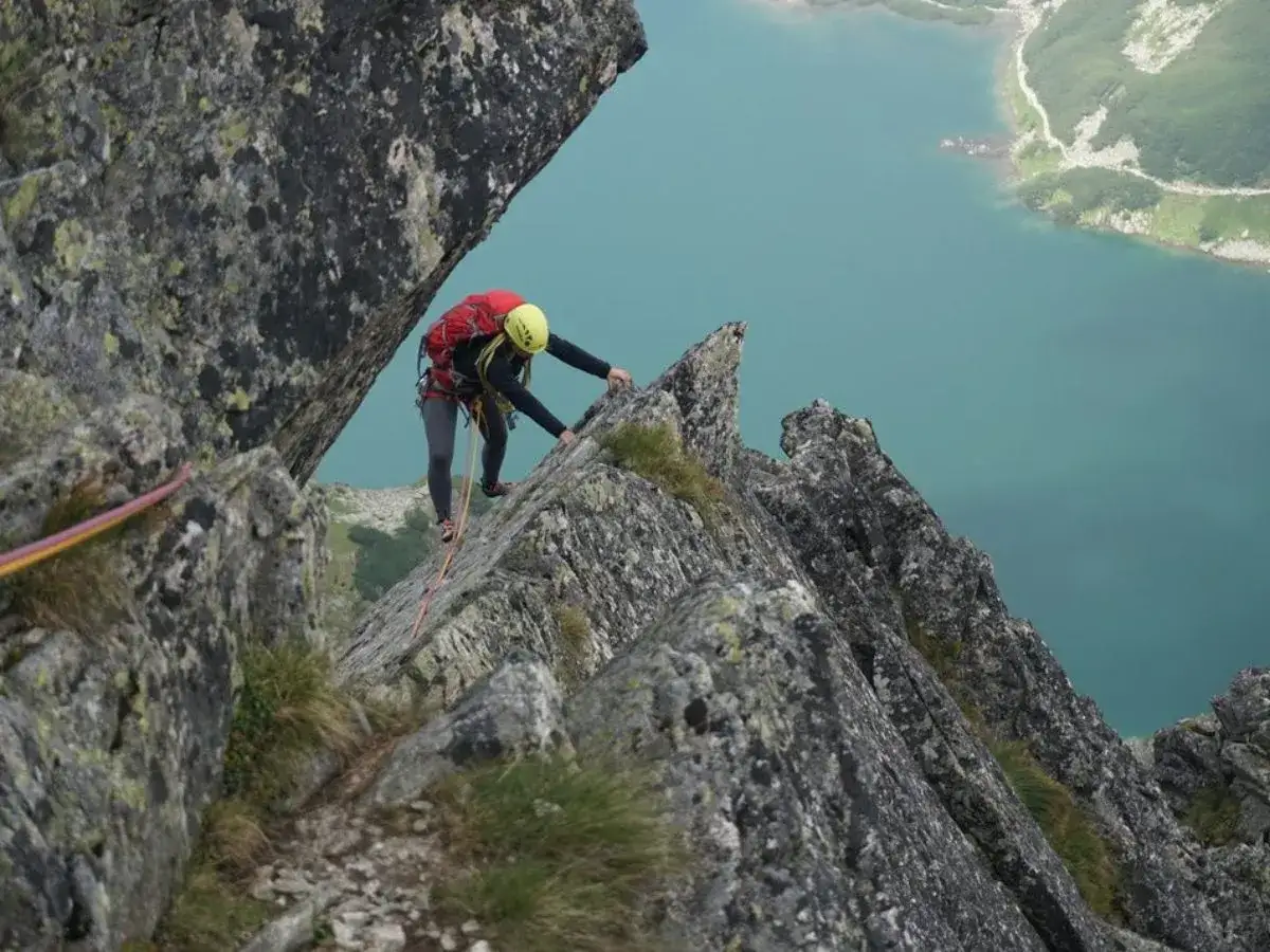 Wspinaczka górska nad turkusowym jeziorem. Alpinistka w kasku i czerwonym plecaku pokonuje skalną grań.