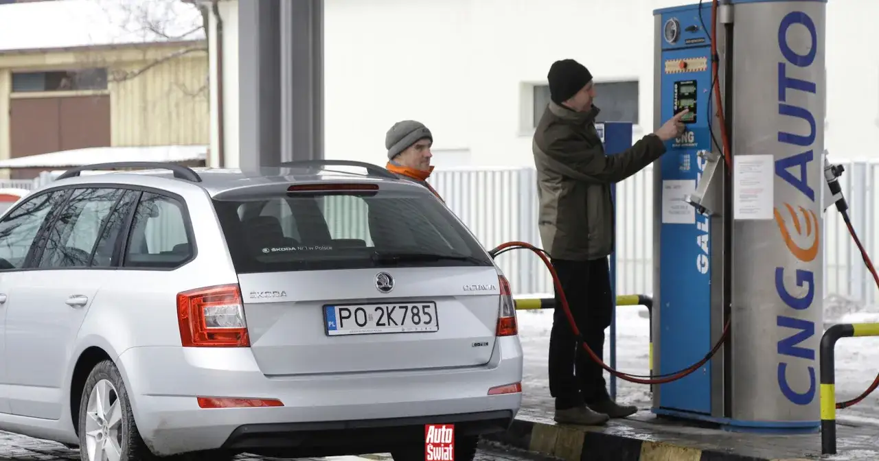 Mężczyzna tankuje gaz CNG do srebrnego kombi na stacji paliw. Obok stoi drugi mężczyzna.