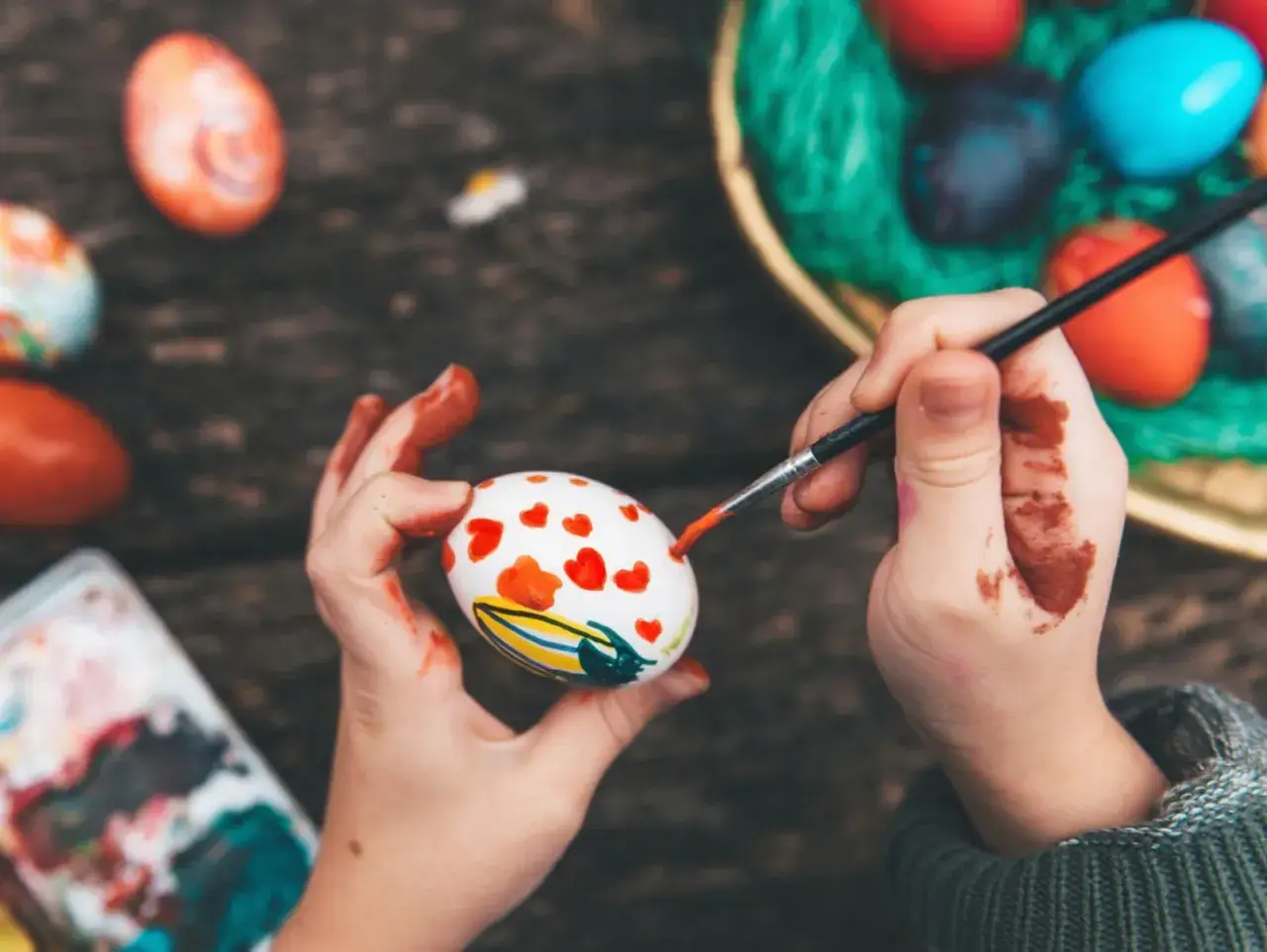 Kind malt mit Pinsel und Farbe bunte Oster Eier. Kleine rote Herzen zieren das Ei, während andere Eier im Korb warten.