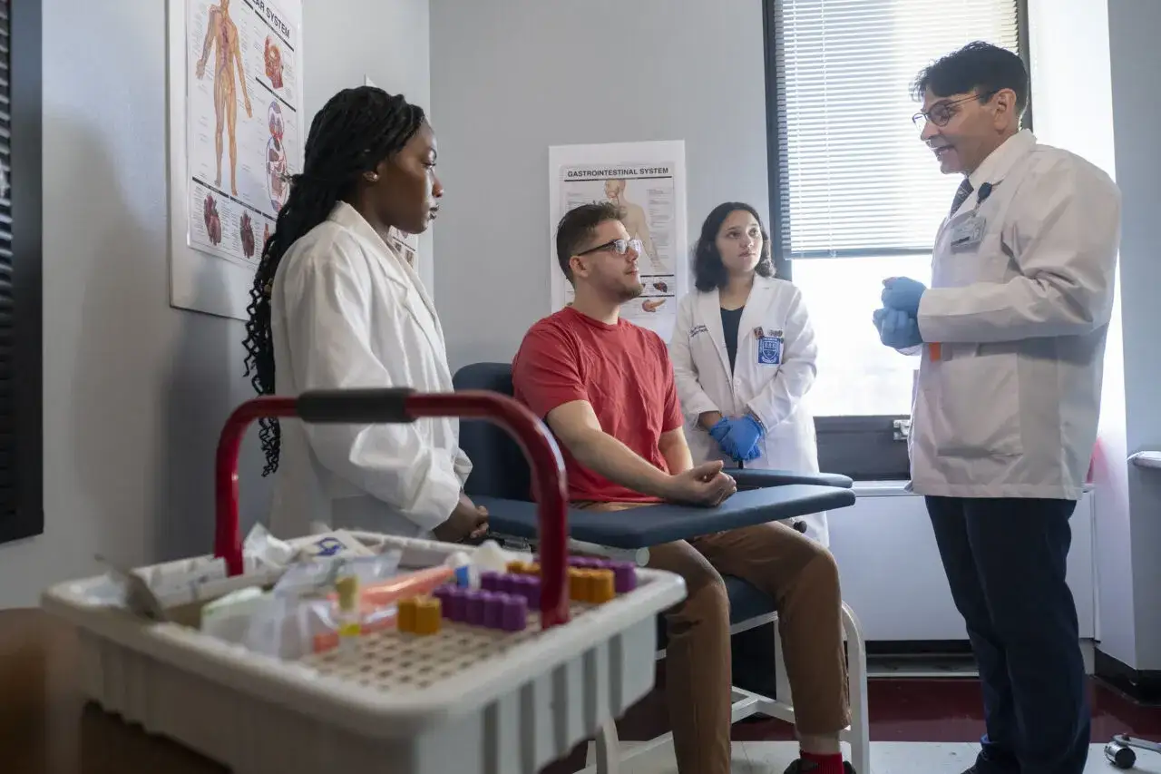 Studenten im Bachelor Gesundheitsmanagement &uuml;ben Blutentnahme. Ein Mann sitzt, zwei Studentinnen und ein Arzt beobachten.