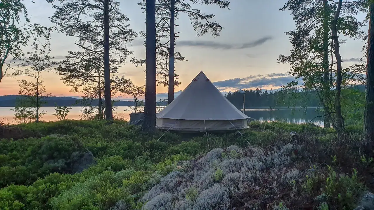 Ett stort tält står på en kulle vid en sjö. Solnedgången färgar himlen i mjuka toner. Perfekt för naturcamping lagom.
