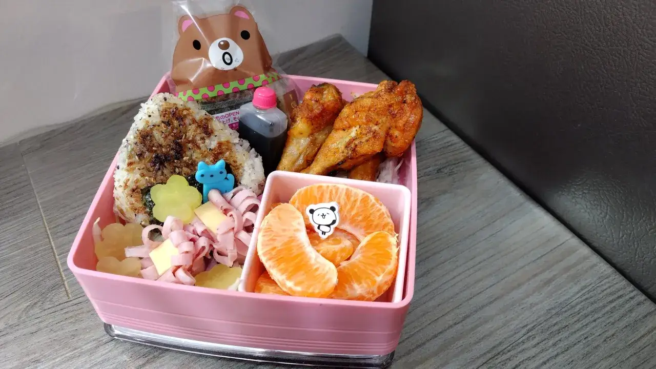 A cute onigiri bento box with chicken wings, mandarin orange slices, ham, and cheese. A bear-shaped snack bag peeks out.
