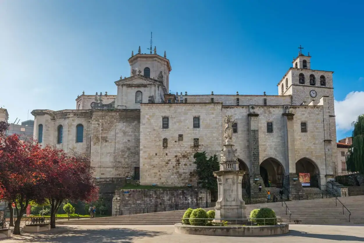 La imponente catedral de Santander se alza bajo un cielo azul, con una estatua en primer plano y un &aacute;rbol de hojas rojas a la izquierda.