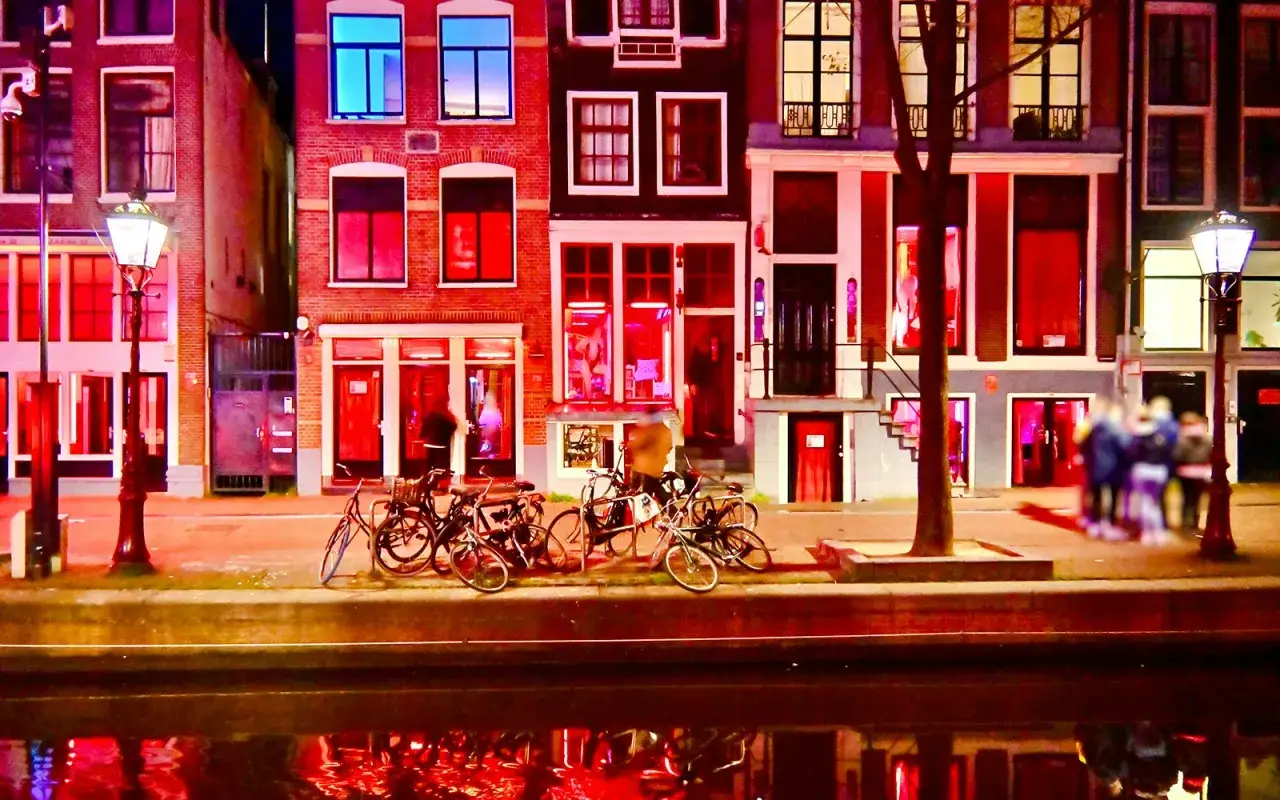 Nocna czerwona ulica Amsterdamu, z oknami w czerwonym świetle i odbiciami w kanale. Rowerzyści zostawili swoje pojazdy, by zanurzyć się w atmosferze.