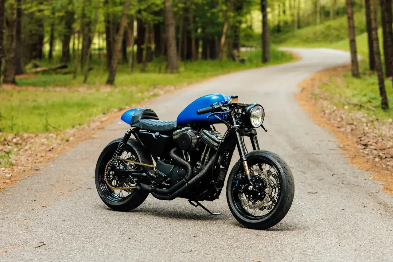 Impresionante **Harley Davidson cafe racer** azul brillante, lista para devorar kil&oacute;metros en un camino boscoso.