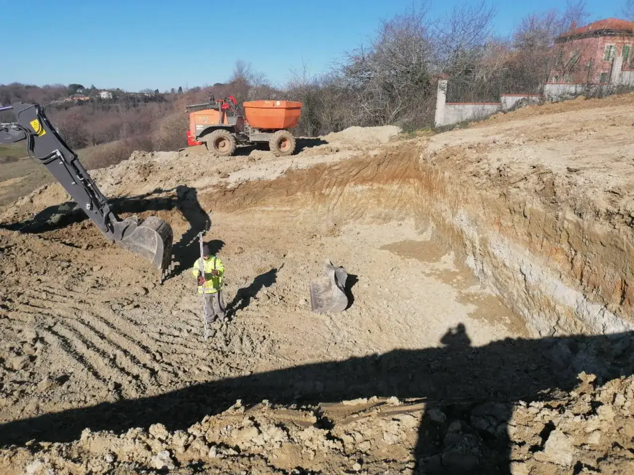 Un ouvrier en jaune v&eacute;rifie les niveaux d'un chantier o&ugrave; une excavatrice et un tombereau sont utilis&eacute;s pour d&eacute;caisser le terrain.