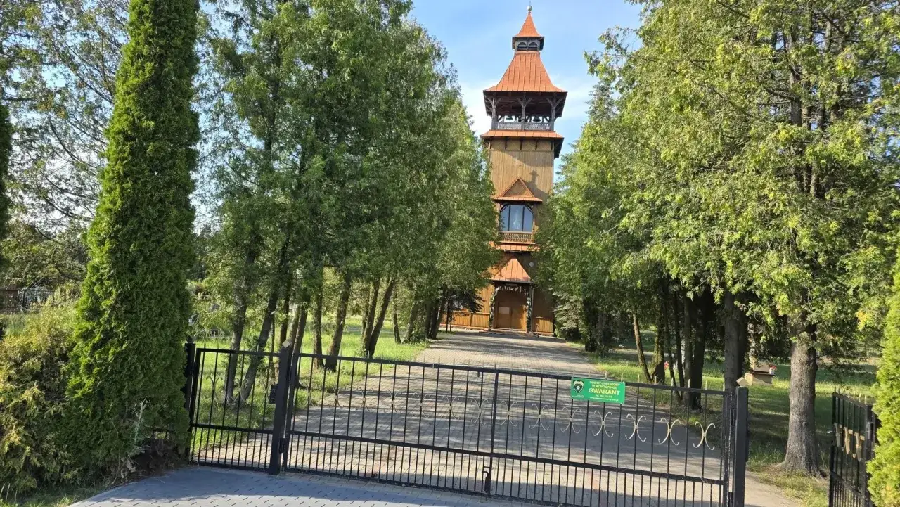 Drewniana wieża w Długosiodle otoczona drzewami, z bramą i znakiem 
