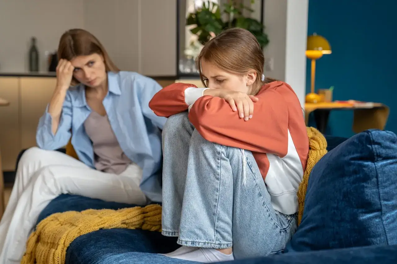 Una figlia femmina si rannicchia sul divano, mentre la madre narcisista la osserva con aria distante e preoccupata.