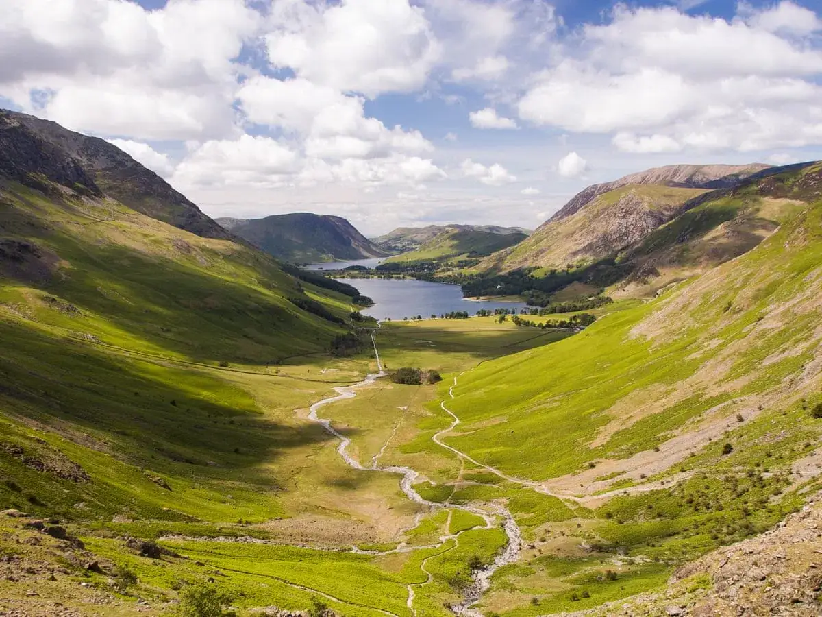 A winding path leads through a lush green valley towards a serene lake, perfect for a c2c hike.