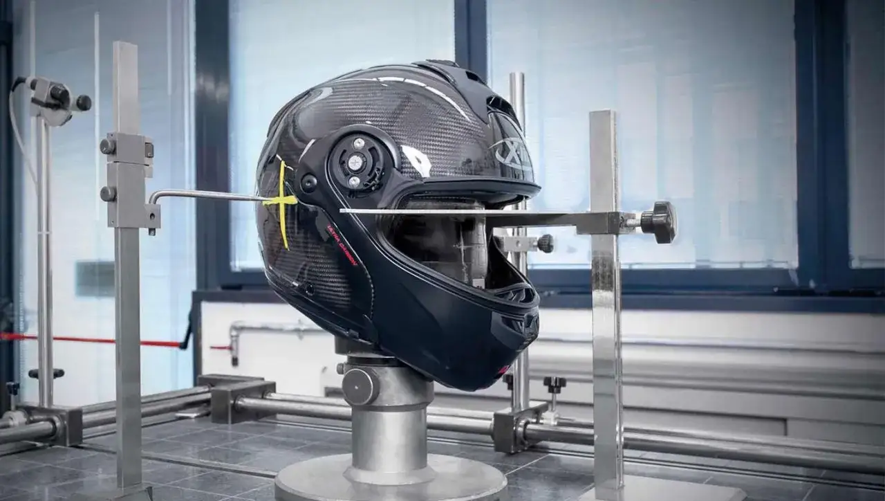 Casco in carbonio sottoposto a test di sicurezza, con un puntale che tocca la visiera. La certificazione Snell E1 garantisce la massima protezione.