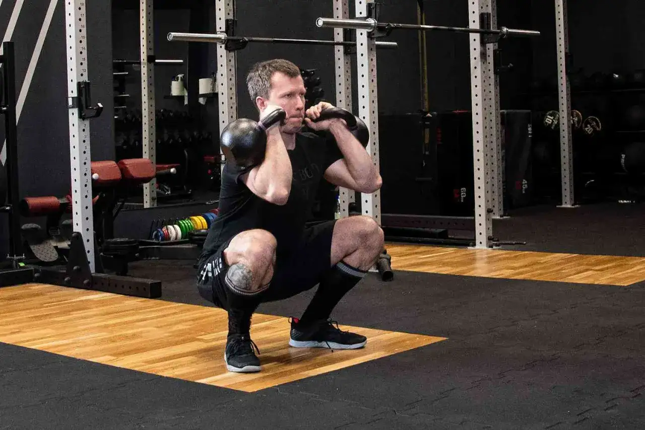 Mężczyzna wykonuje przysiad z dwoma kettlebellami, ćwiczenie typowe dla crossfitu.