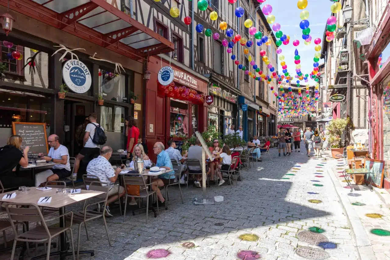 Terrasse animée dans une rue pavée de Limoges, décorée de guirlandes de ballons colorés. Des clients dégustent un repas, profitant de l'ambiance estivale. Une enseigne